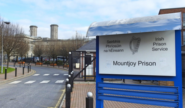 The towers of Mountjoy prison are in the background. In the foreground is the entranceway to the prison with gates and a gapekeeper's hut. A sign reads: Mountjoy Prison 