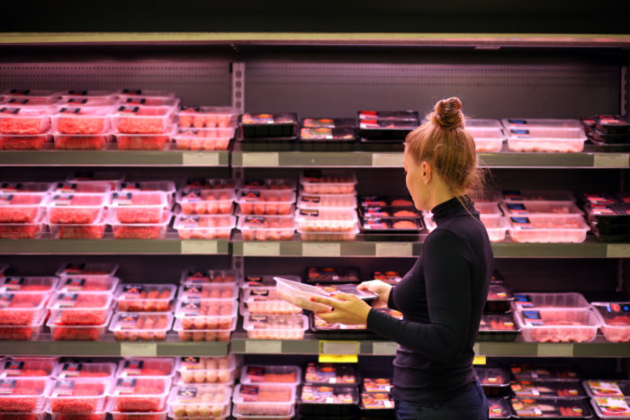 Women holding two packets of meat in front of a meat section in a supermarket.