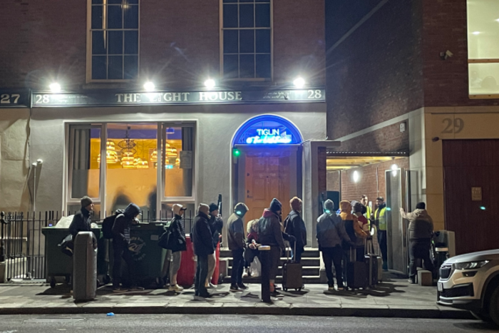 A queue of over 10 people, wearing winter clothes, many with suitcases and bags, outside the cafe