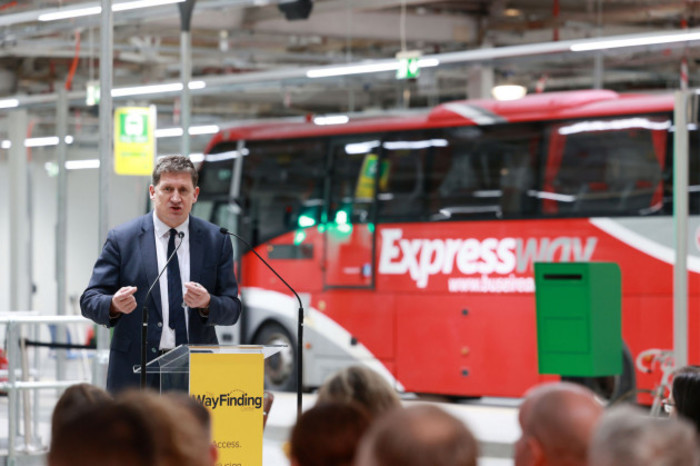 Ryan wearing a suit and speaking at a podium in front of a group of people. A red bus with 'Expressway' written on it is behind him.